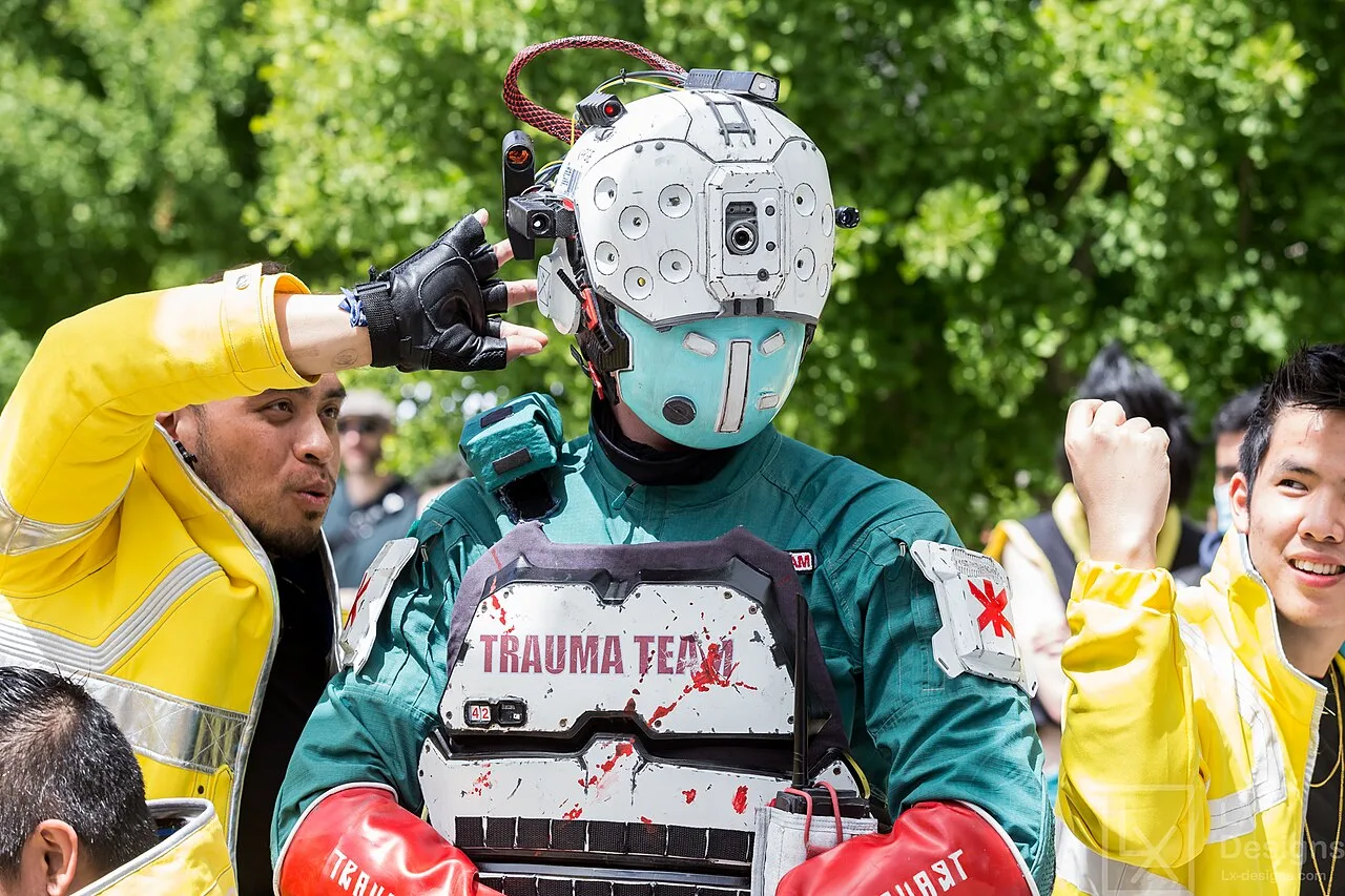 Косплеер в форме Trauma Team из Cyberpunk 2077: белый шлем с камерами, бронежилет с пятнами крови, рядом фанаты в жёлтом